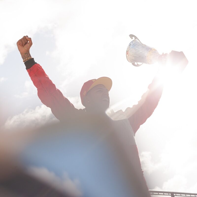 Pilote de course en combinaison rouge levant les bras au ciel avec un trophée, symbolisant la victoire finale rendue possible par un accompagnement fidèle.