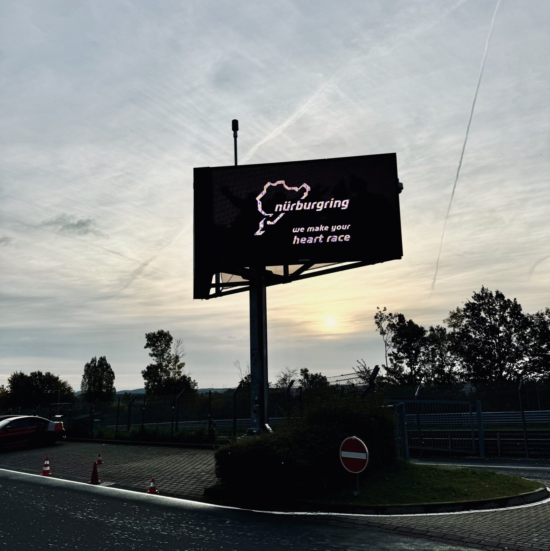Grand panneau d'affichage numérique noir à l'entrée du circuit du Nürburgring affichant le tracé de la Nordschleife et le slogan "we make your heart race" au coucher du soleil.